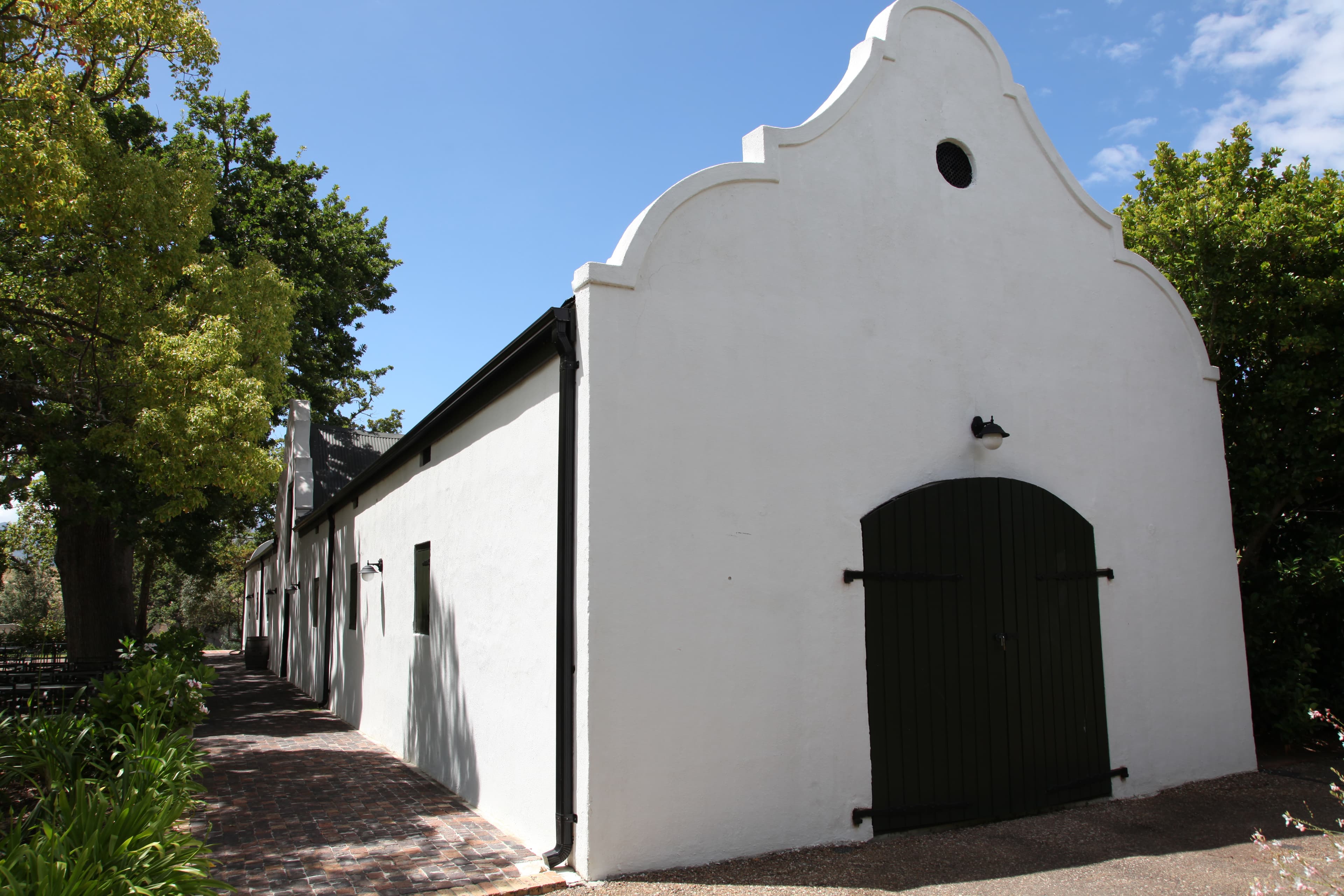 The white holbol-gabled old wine cellar at Spier, dated 1767 and the estate's oldest building, seen along a brick-paved lane beneath oak trees.